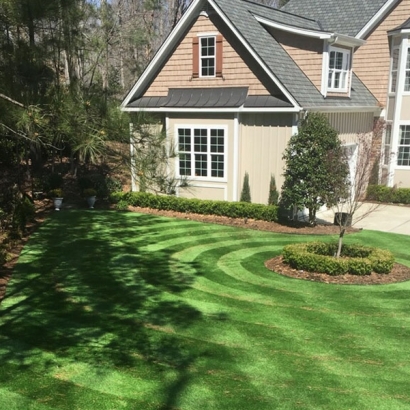 Front yard landscape with striped Global Syn-Turf Spring-50 artificial grass, round flower bed, two-story house.